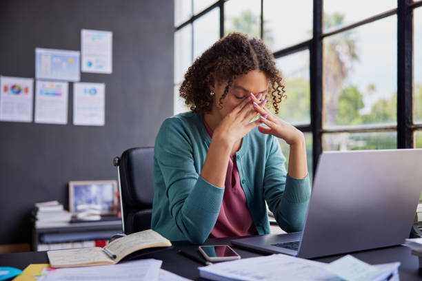 Woman sitting at a desk with a laptop, rubbing her eyes in frustration among scattered papers and notes.