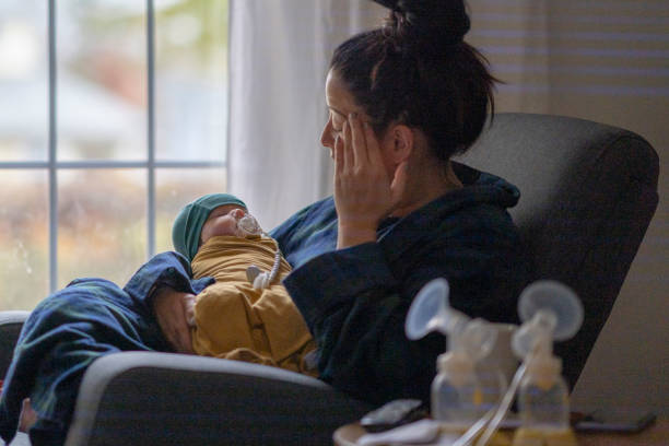A tired mother sits in a chair and cradles her newborn wrapped in mustard yellow clothing, by a window with a hand resting on her face.